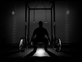 Full length of silhouette teenage boy holding barbell in darkroom