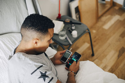 High angle view of man holding glaucometer checking mobile phone while lying on bed at home