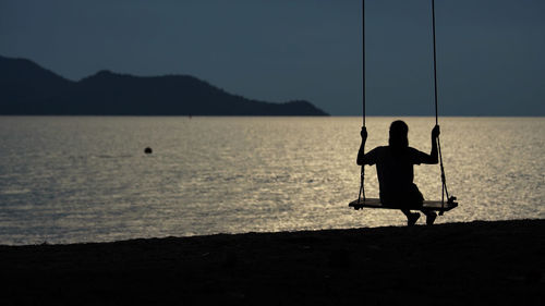 Rear view of silhouette woman sitting on swing at beach against clear sky