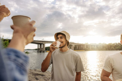 Smiling young man wearing bucket hat enjoying drink with friend during picnic