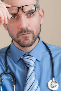 Portrait of doctor holding eyeglasses while wearing stethoscope against wall