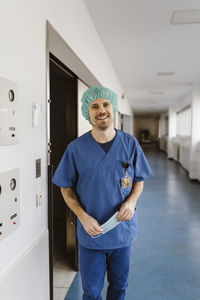 Portrait of smiling male physician wearing scrubs standing at hospital corridor