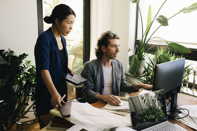 Female architect holding wooden plank while standing by male colleague using computer