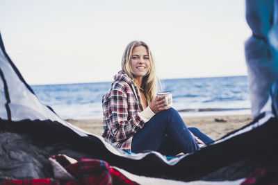 Portrait of young woman camping at beach against clear sky