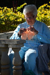 Woman knitting while sitting on seat against plants