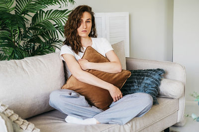 A sad woman sits on a sofa at home hugging a pillow.