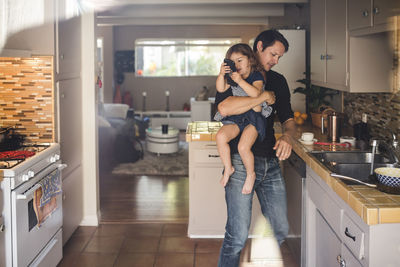 Daughter using phone while father carrying and working in kitchen