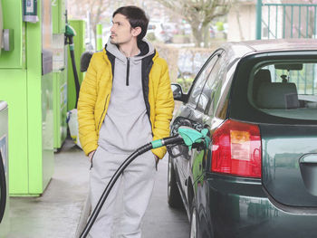 Handsome young caucasian man in yellow jacket and gray sport refuels his car