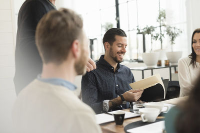 Happy businessman reading document while sitting with colleagues at table in office