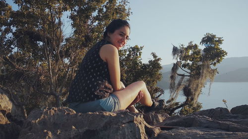 Portrait of smiling woman sitting on rock by lake 