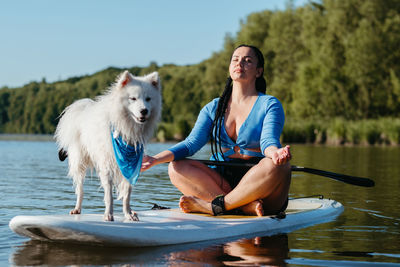Young woman meditating on the city lake while sitting on the sup board with her dog japanese spitz
