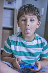 Portrait of boy sticking out tongue while playing handheld video game at home