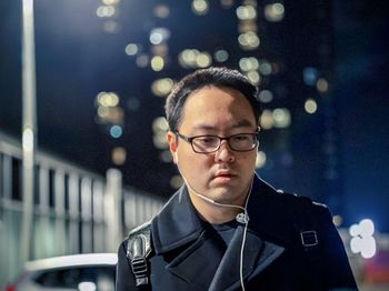 Young asian man in eyeglasses and earphones standing on city street at night with illumination.
