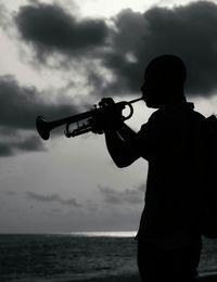 Side view of silhouette man playing trumpet by sea against cloudy sky