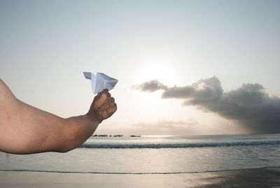 Cropped hand of man flying paper airplane at beach against sky during sunset