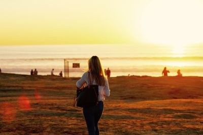 Rear view of woman with shoulder bag walking at beach against sky during sunset