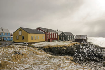 Houses on a cliff, painted in primary colors under a dark sky on which the sun breaks through