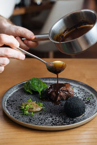 Cropped anonymous person hands pouring sauce on delicious gourmet meat steak with sauce and herbs served on metal silver plate