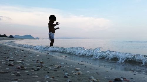 Side view of boy standing on shore at beach against sky during sunset