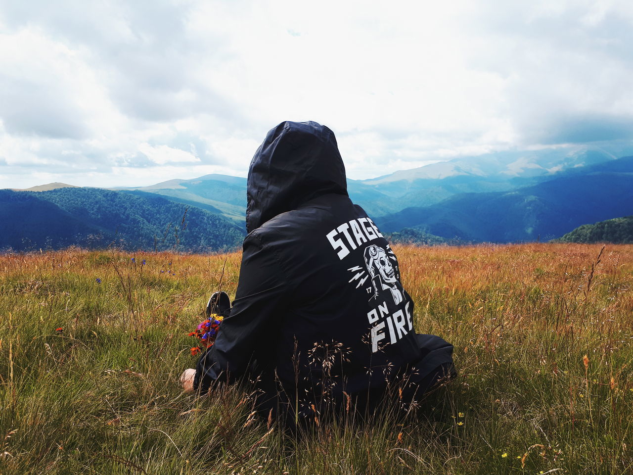 REAR VIEW OF WOMAN SITTING ON FIELD AGAINST MOUNTAIN