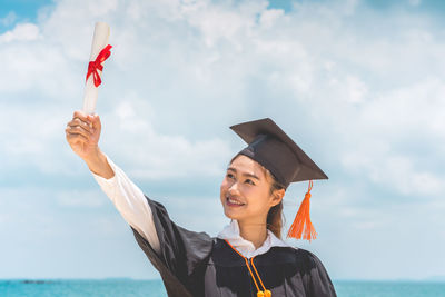 Smiling woman in graduation gown holding diploma while standing against sea