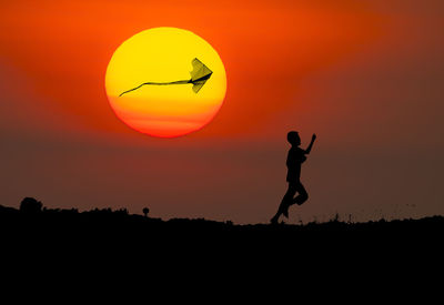 Silhouette boy flying kite while running on field against sun during sunset