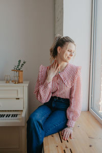 A pretty young woman in jeans and a plaid blouse sits on the windowsill by the window.