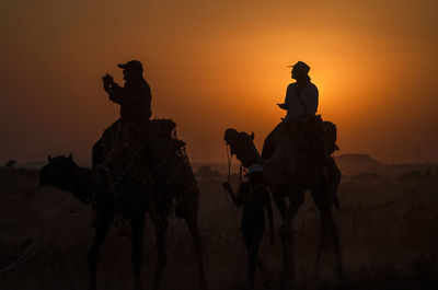 Silhouette tourists sitting on camels against clear orange sky during sunset
