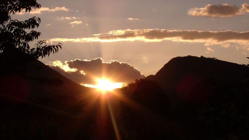 Sunlight streaming through silhouette trees against sky during sunset