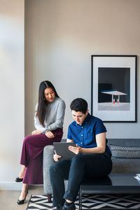 Male and female colleagues discussing over graphics tablet while sitting on sofa against wall at office