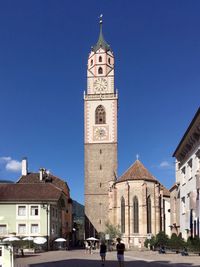 People on street leading towards clock tower and buildings against blue sky