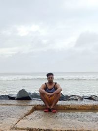 Young man sitting on shore at sea against sky