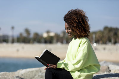 Side view of smiling woman with curly hair reading book while sitting at beach
