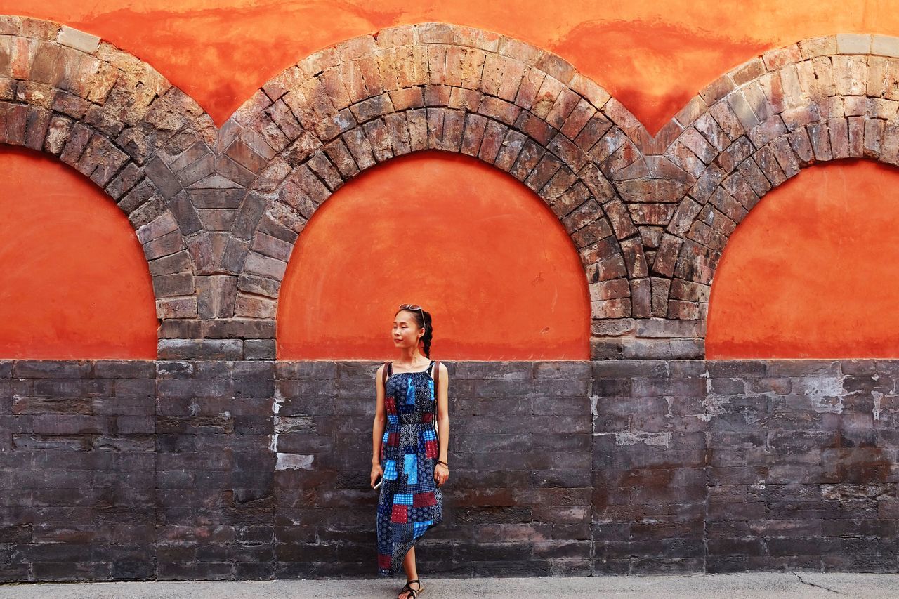 lifestyles, casual clothing, standing, leisure activity, full length, built structure, architecture, young adult, person, building exterior, front view, young women, brick wall, wall - building feature, portrait, red, looking at camera, smiling
