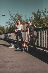 Young women with dogs standing on footbridge against clear sky during sunny day