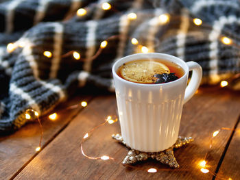 White cup of hot tea with lemon. cozy decorations with grey knitted sweater and light bulbs.