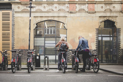 Full length rear view of couple renting bicycles from bike share stand in city