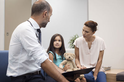 Mother and doctor looking at girl while sitting on sofa in hospital