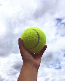 Cropped hand holding ball against cloudy sky