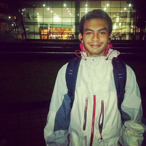 PORTRAIT OF SMILING YOUNG MAN STANDING AGAINST ILLUMINATED WALL