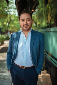 Happy hispanic mature male manager in suit holding hands in pockets and looking at camera with smile while leaning on glass wall on sunny day on city street