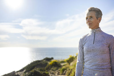 Senior woman looking away while standing on cliff by sea against sky during sunny day