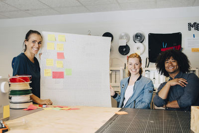 Portrait of smiling multi-ethnic technicians with adhesive notes on pegboard at creative office