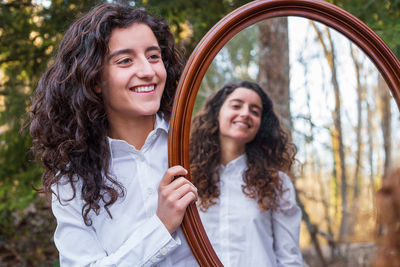 Smiling woman holding mirror with sister reflection at forest