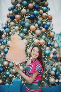 Portrait of smiling young woman standing by christmas tree holding cotton candy outdoors