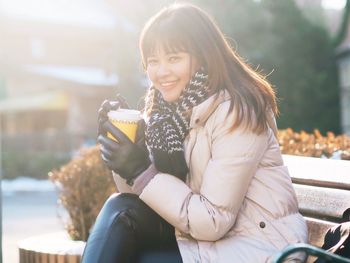 Portrait of young woman holding disposable cup while sitting on bench during winter