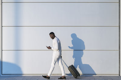 Man with suitcase using smart phone while walking by white wall