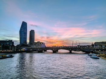 Bridge over river by buildings against sky during sunset