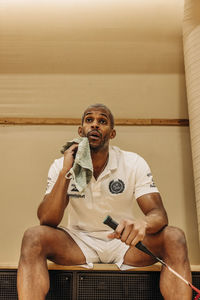 Tired male badminton player wiping sweat with towel sitting at sports court