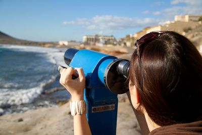 Rear view of woman looking through binoculars at beach against sky